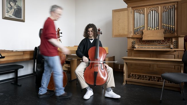 Person spielt Cello, während ein Lehrer daneben steht. Im Raum steht eine große Orgel.