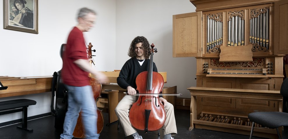 Person spielt Cello, während ein Lehrer daneben steht. Im Raum steht eine große Orgel.