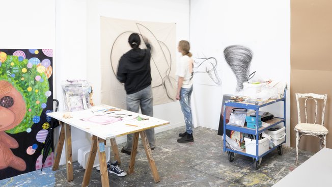 Two people drawing in an art studio, surrounded by art materials and paintings.