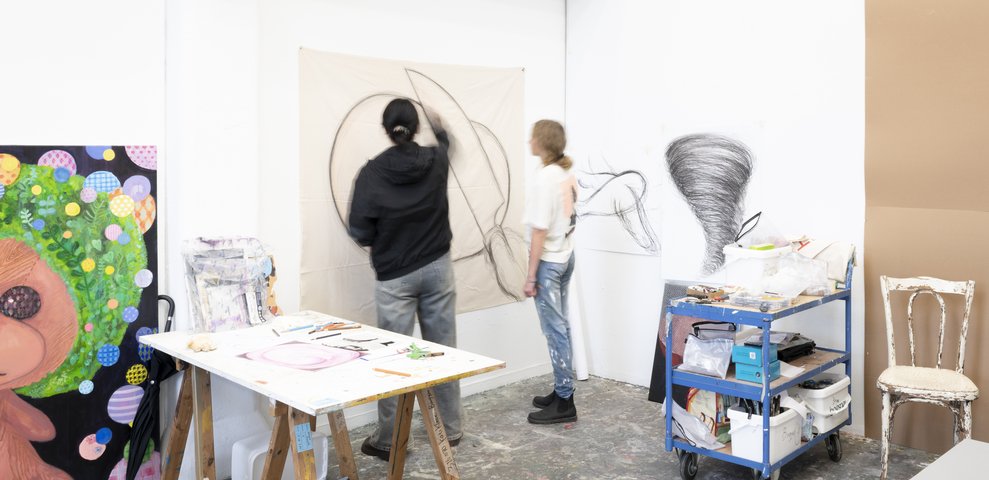 Two people drawing in an art studio, surrounded by art materials and paintings.